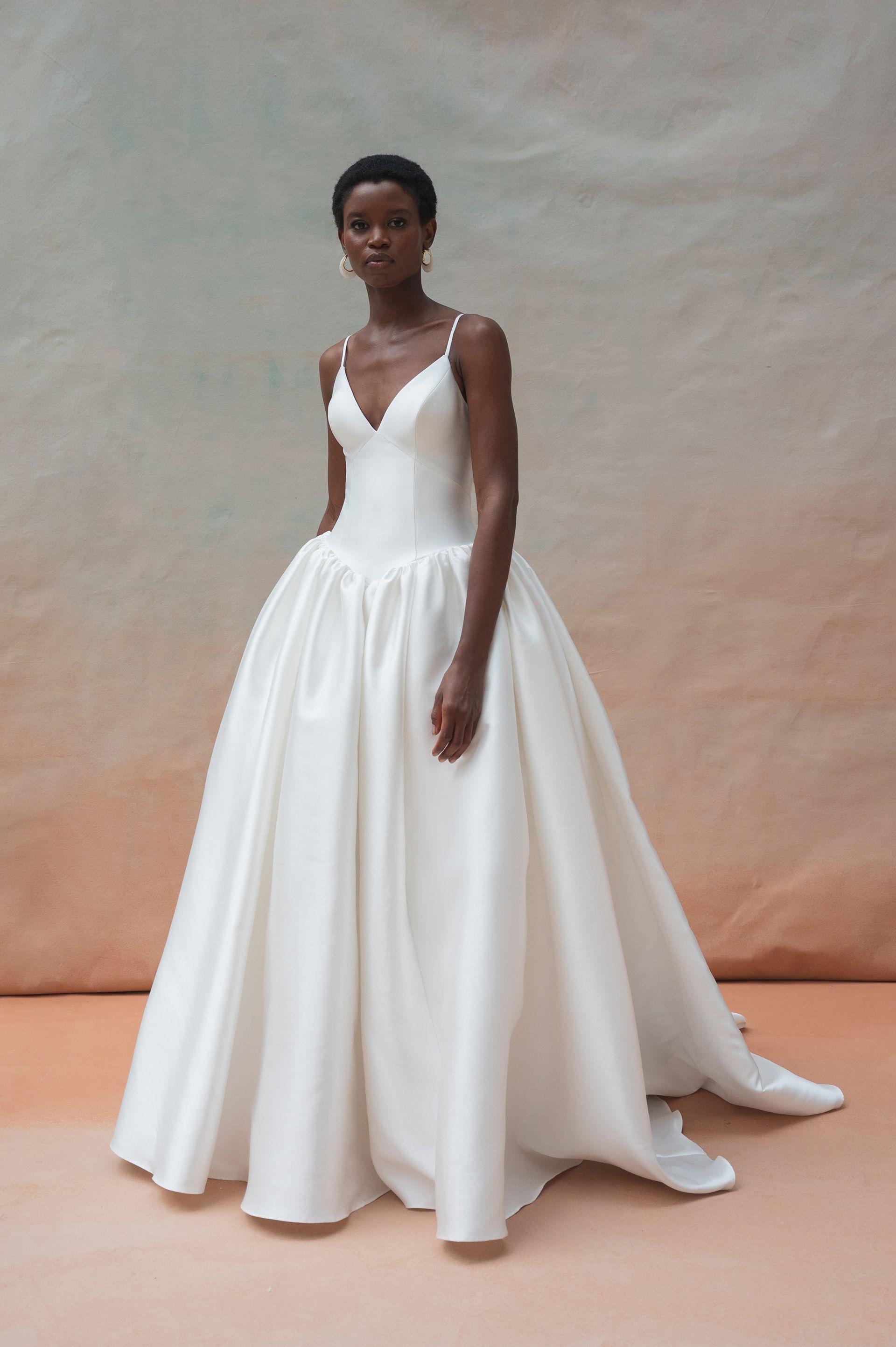 A person poses in a floor-length, white satin ballgown with a V-neckline and thin straps against a neutral backdrop.