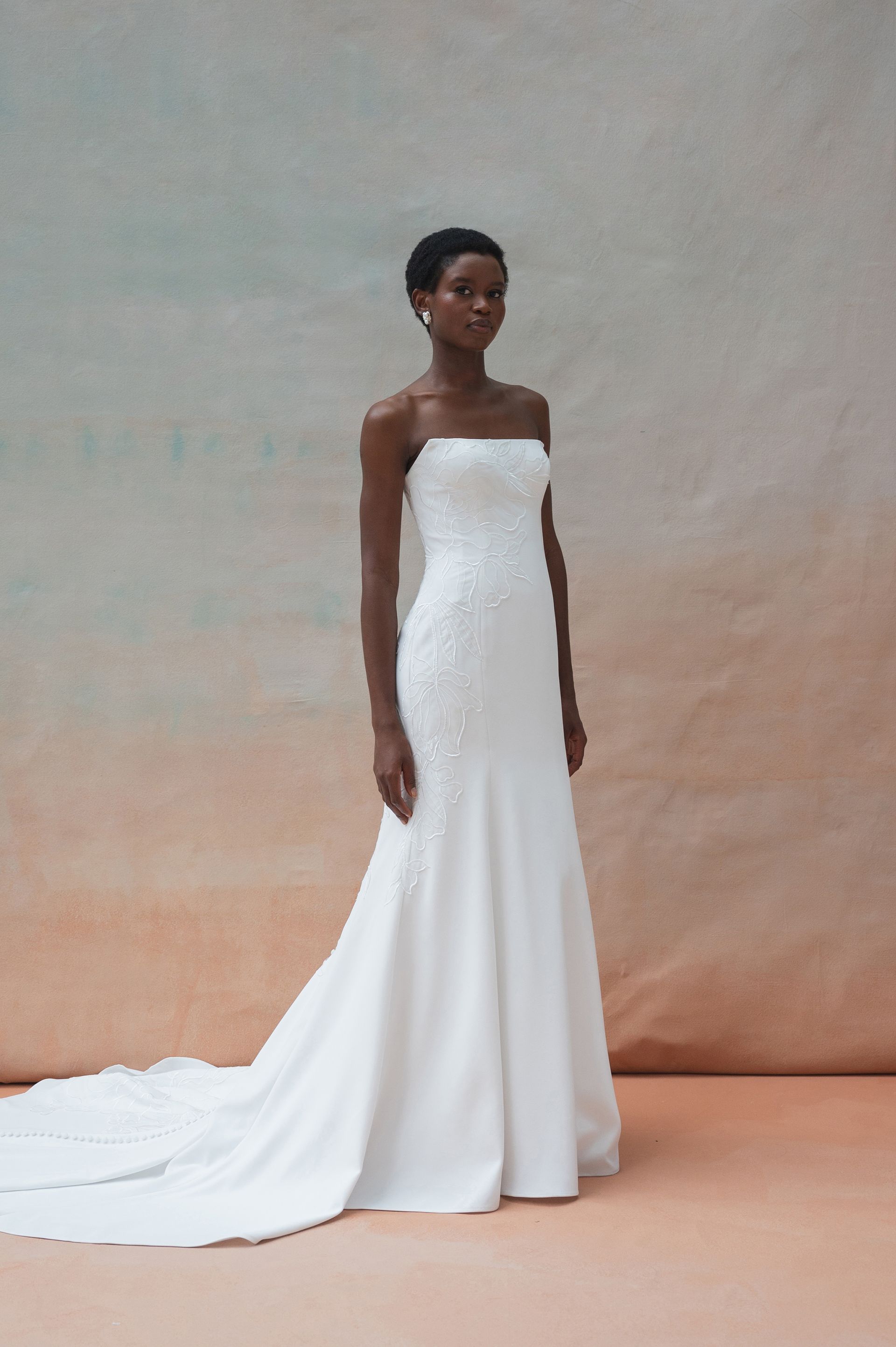 A person wears a strapless white column wedding gown with a train against a neutral, textured backdrop.