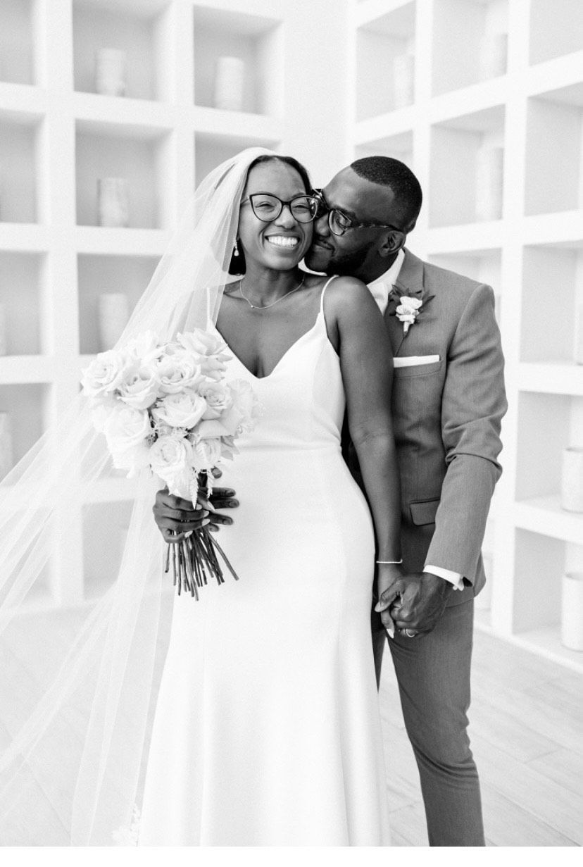 A couple smiles while embracing in wedding attire, standing before a wall with recessed shelves.