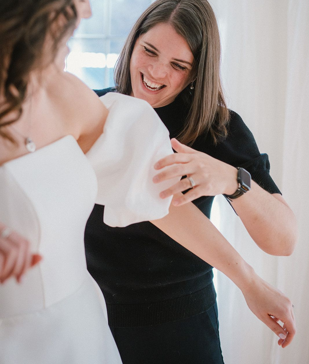 A person in a black top helps a bride adjust the sleeve of her white wedding dress while smiling.