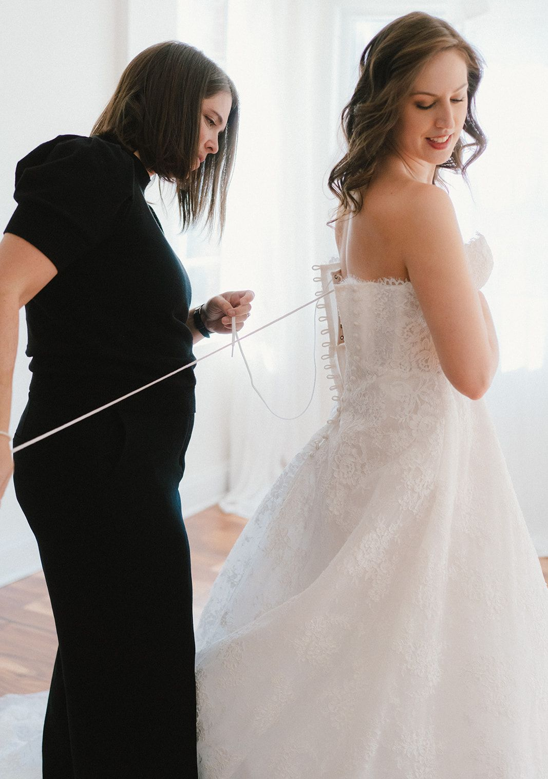 A person helps a smiling bride lace up the back of her white, strapless, lace wedding gown in a bright room.
