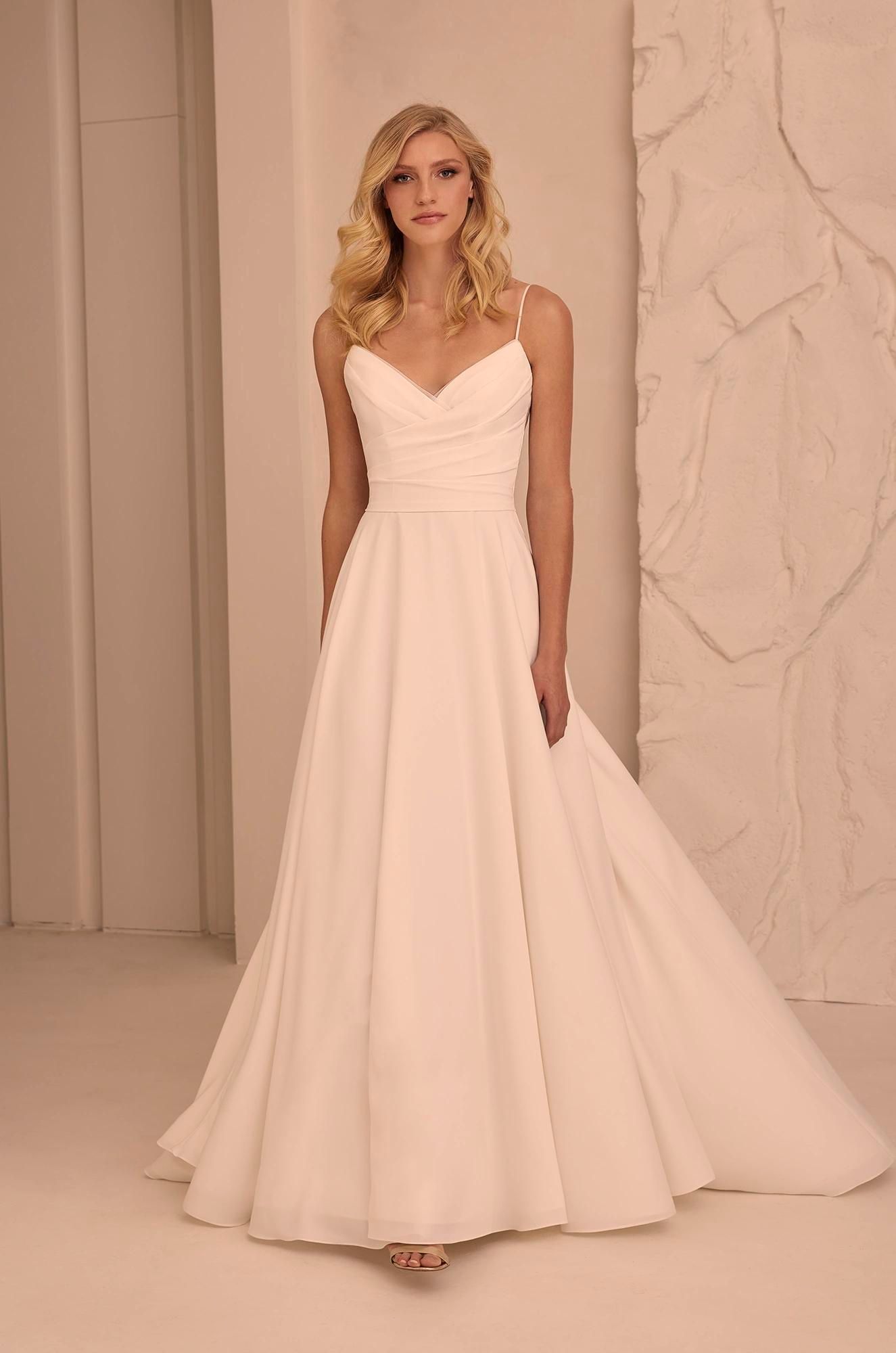 A person stands in a studio wearing a floor-length, pale pink, A-line bridal gown with a draped bodice and thin straps.