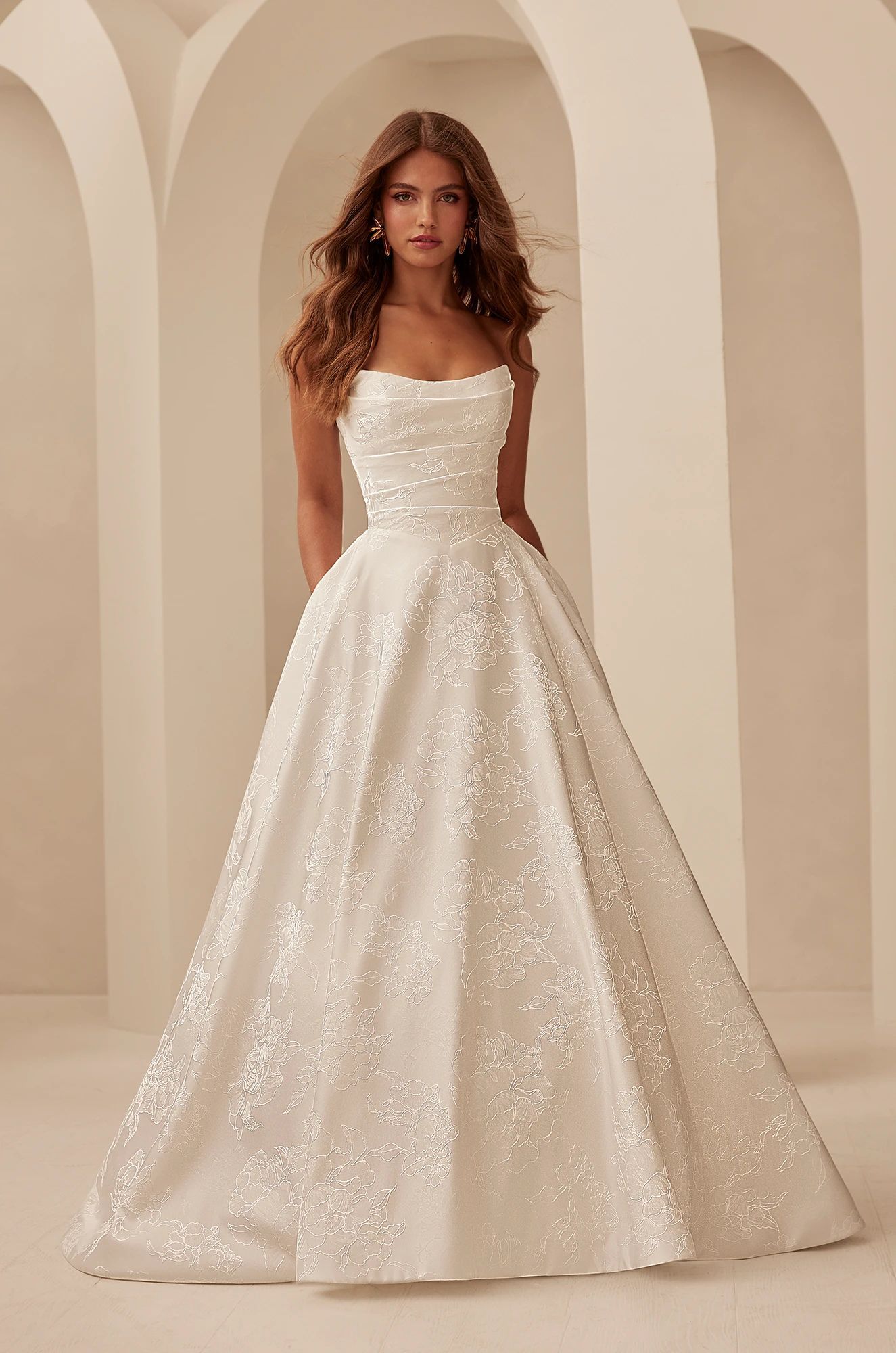 A model wears a strapless, white, A-line ballgown covered in delicate floral lace standing in a neutral archway.