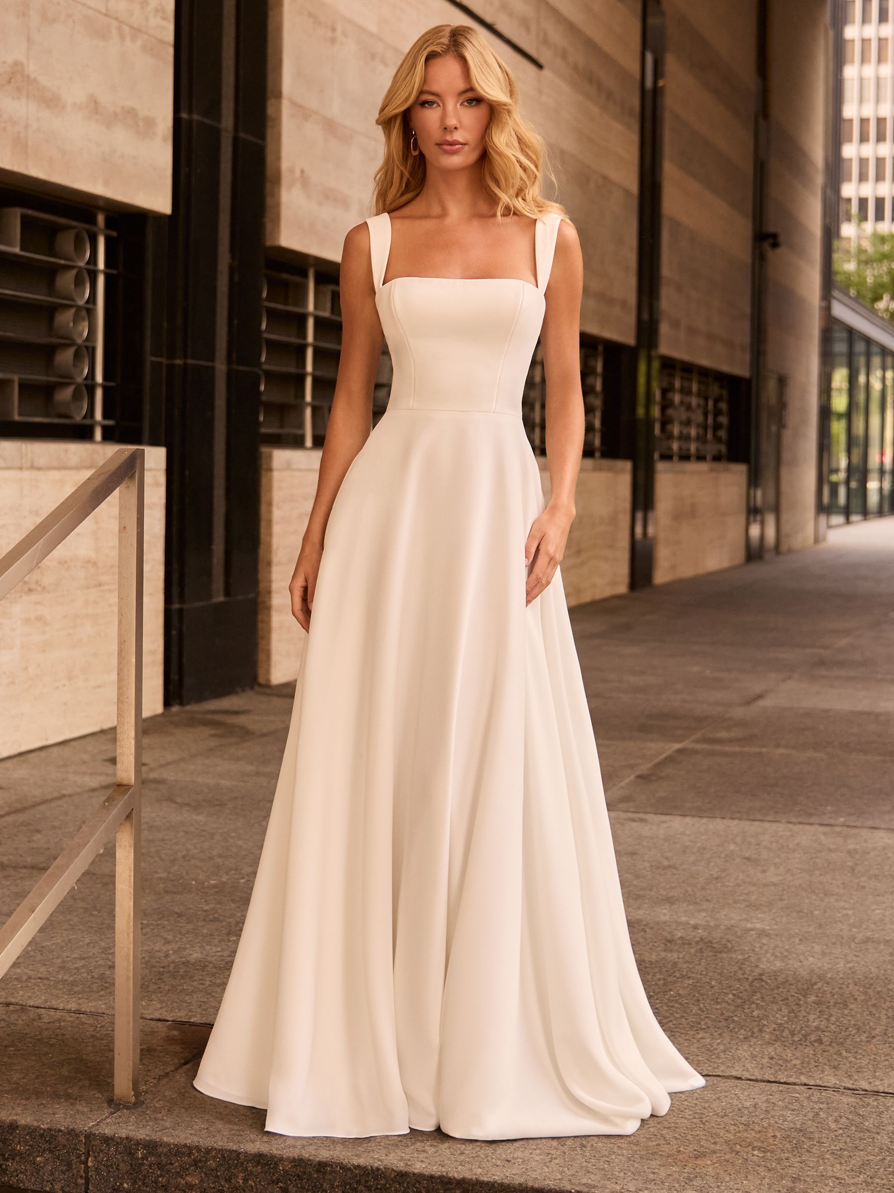 A woman stands outdoors wearing a floor-length, off-white sleeveless gown with a square neckline and a flowing A-line skirt.