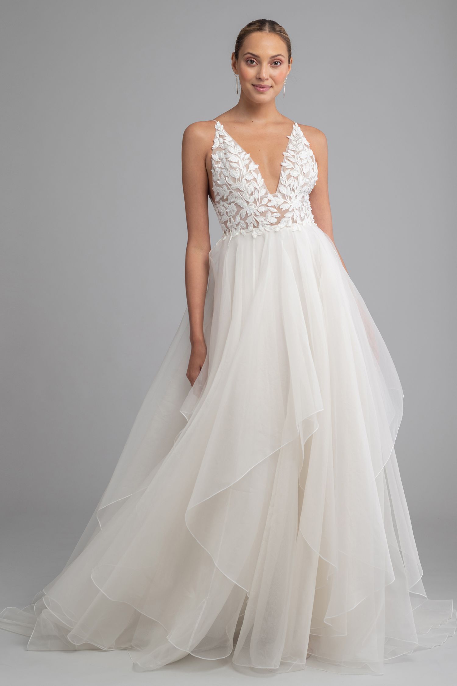 A model wearing a white, sleeveless bridal gown with a deep V-neck, floral lace bodice, and layered, flowing skirt.