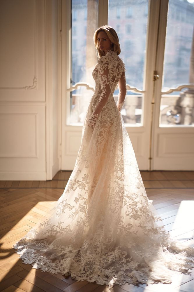A woman stands in a bright room wearing a long-sleeved white lace wedding dress with a full, flowing train.
