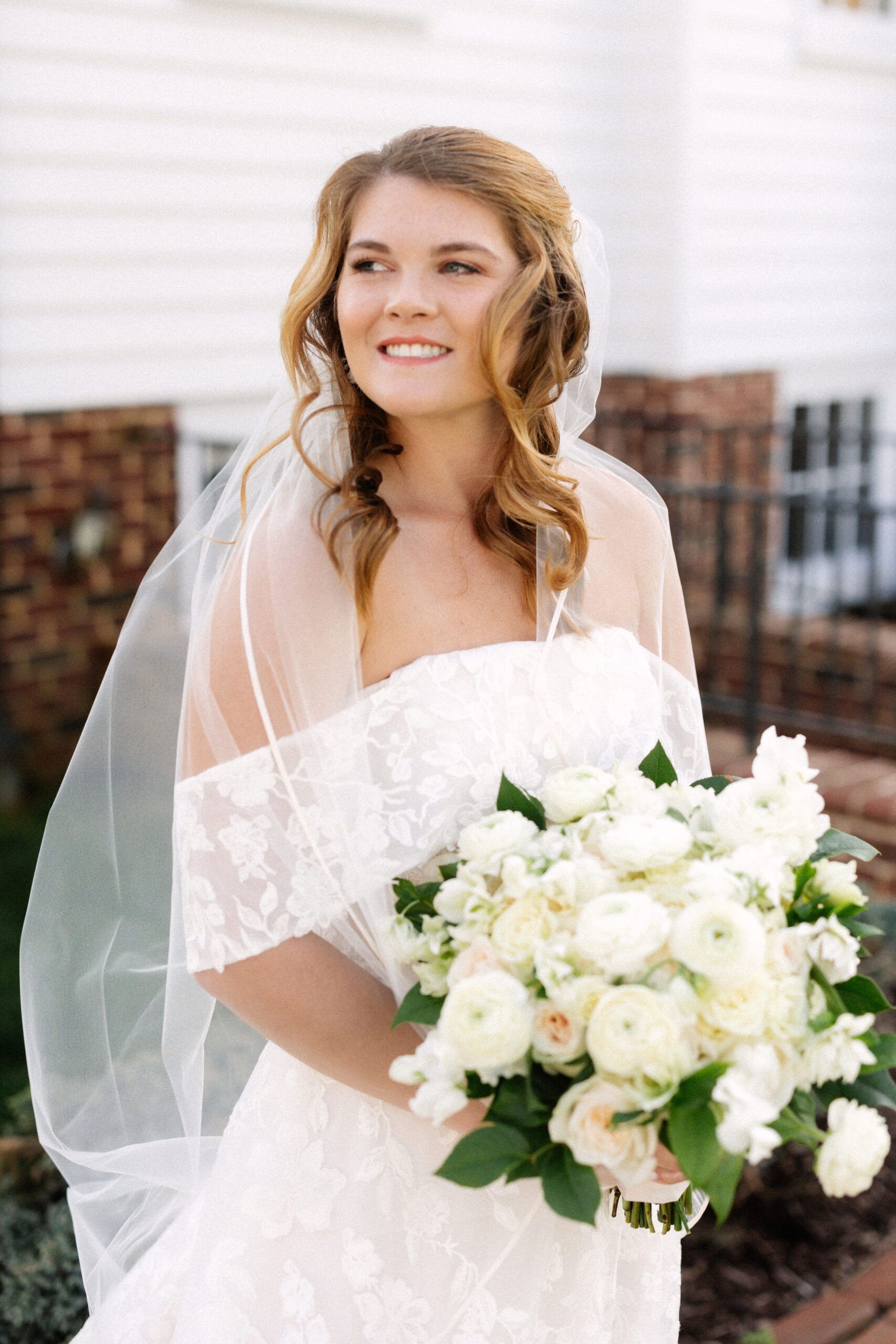 A bride in a white lace off-the-shoulder dress and veil smiles, holding a large bouquet of white flowers outside.