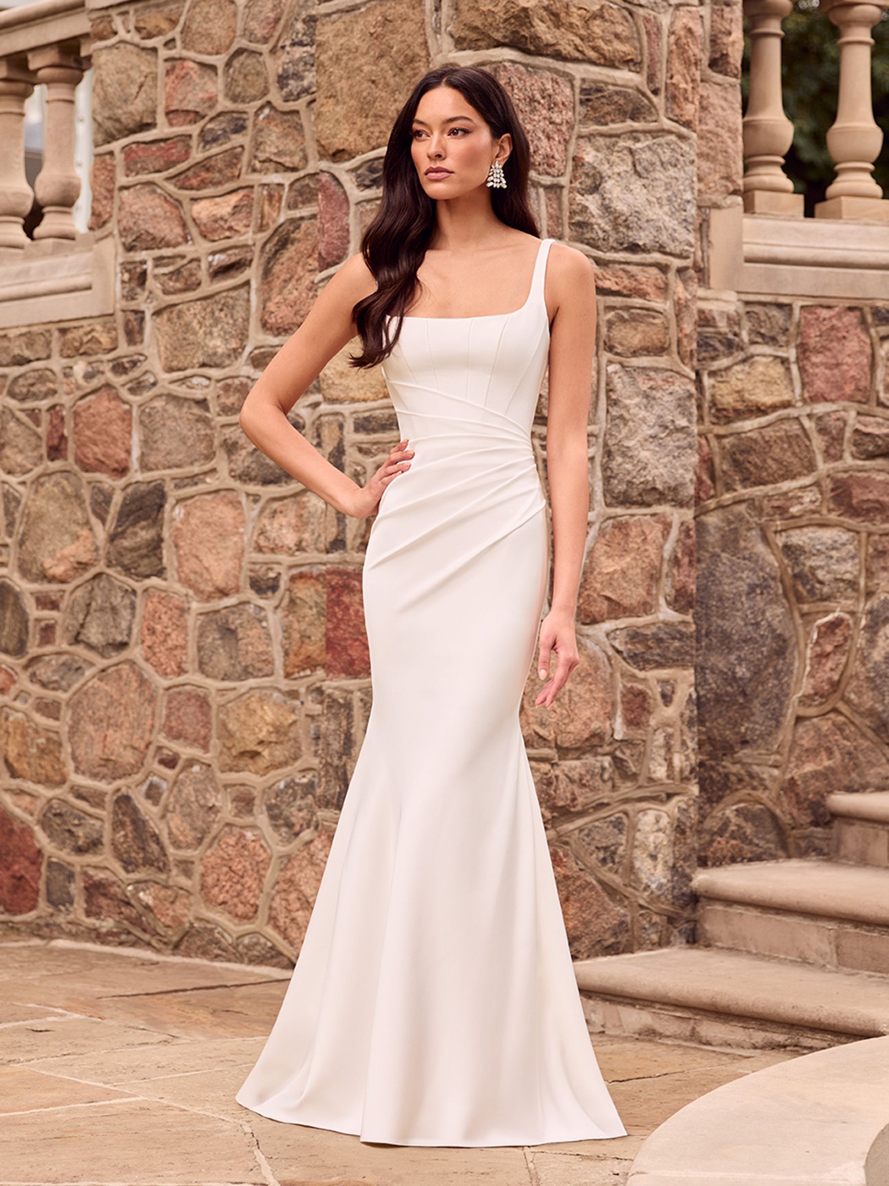 A person models a white, floor-length, square-neck bridal gown with subtle side ruching against a stone-walled background.