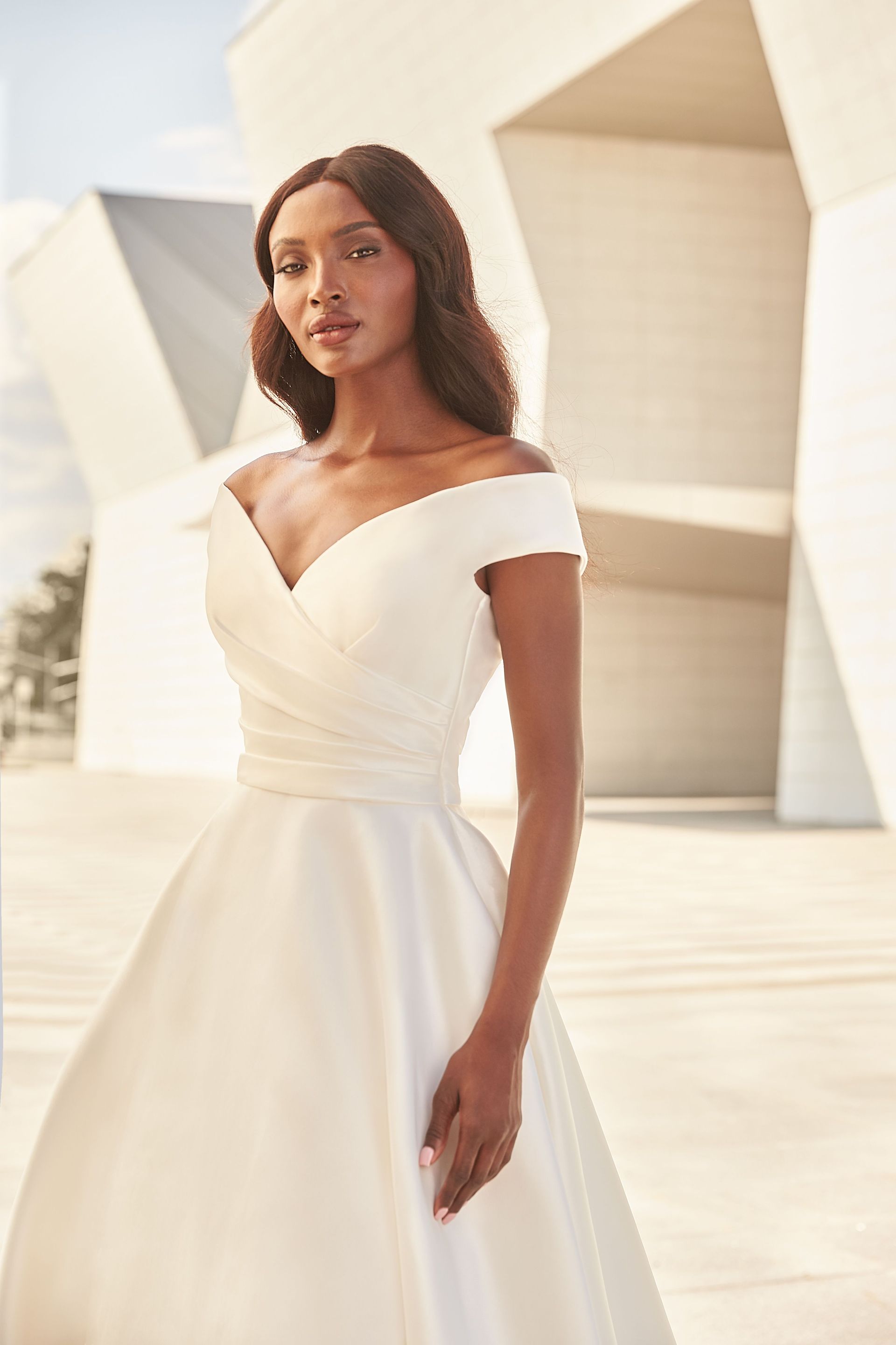 A person wearing an off-the-shoulder, white satin wedding dress with a ruched bodice standing in front of modern structures.
