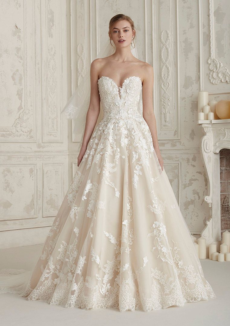A person wearing a strapless, A-line wedding dress with intricate white floral lace over a beige lining in a studio.