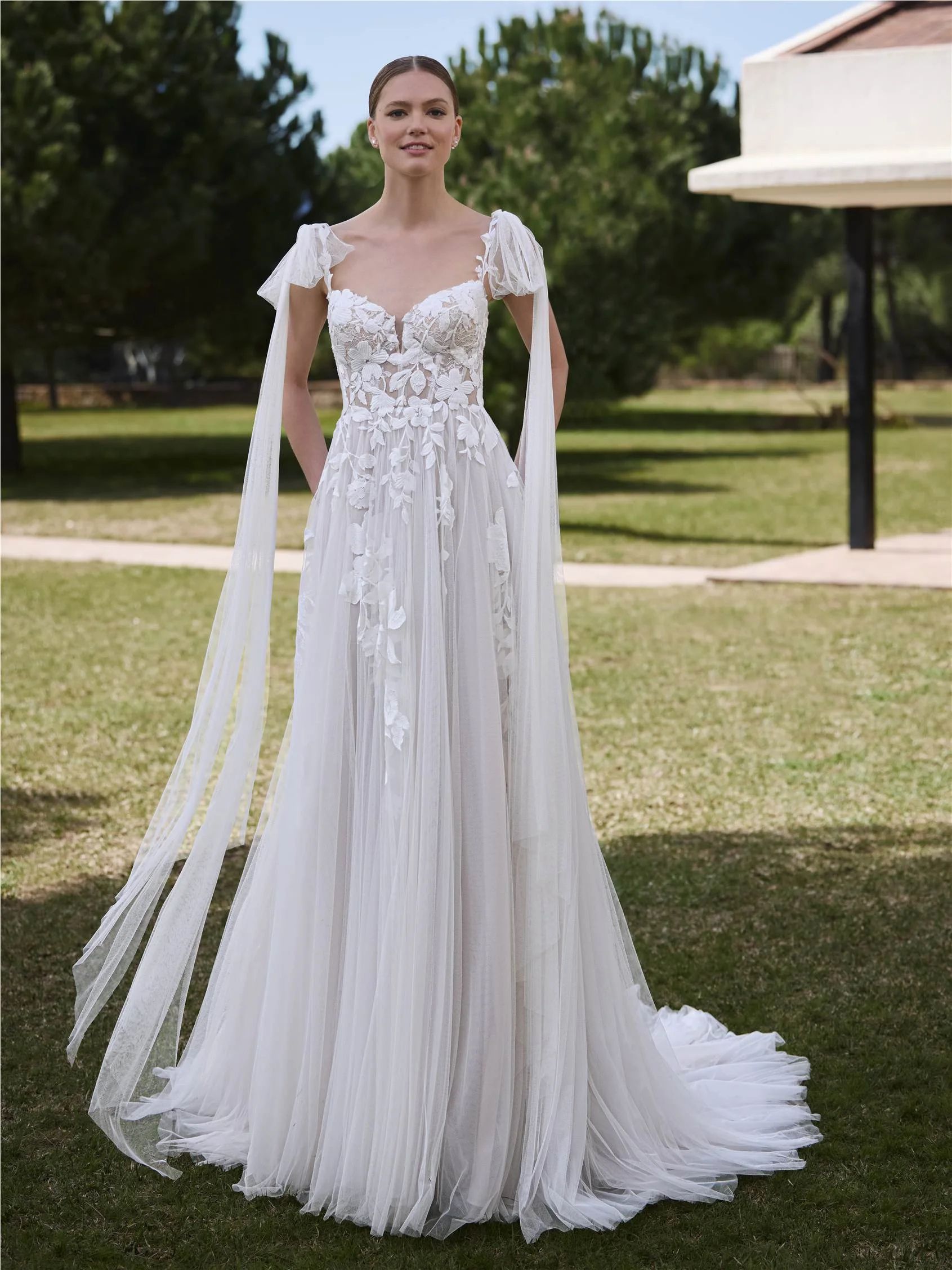 A person standing on a lawn wearing a white, floor-length lace wedding dress with a sweetheart neckline and long sleeves.