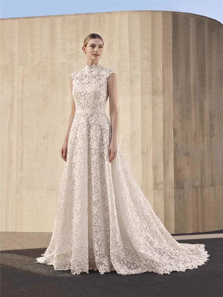 A model wearing a floor-length, high-neck white lace wedding dress with a train, standing before a beige fluted wall.
