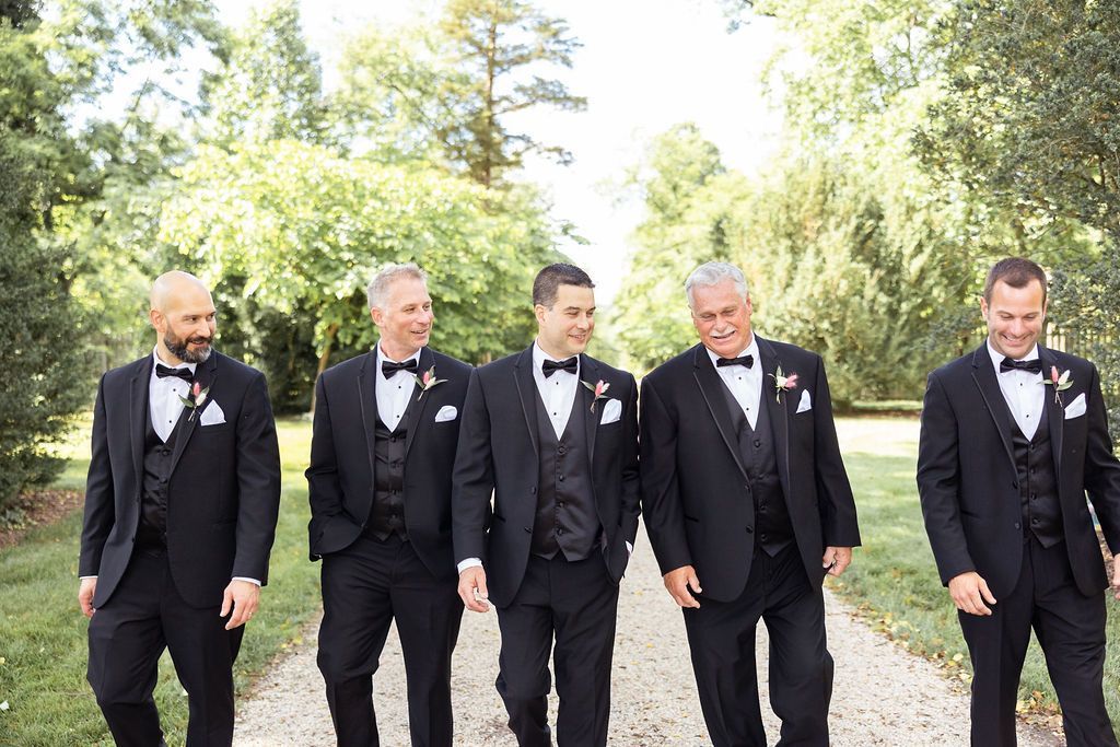 Five men in black suits and ties walking side-by-side on a gravel path outdoors among trees.