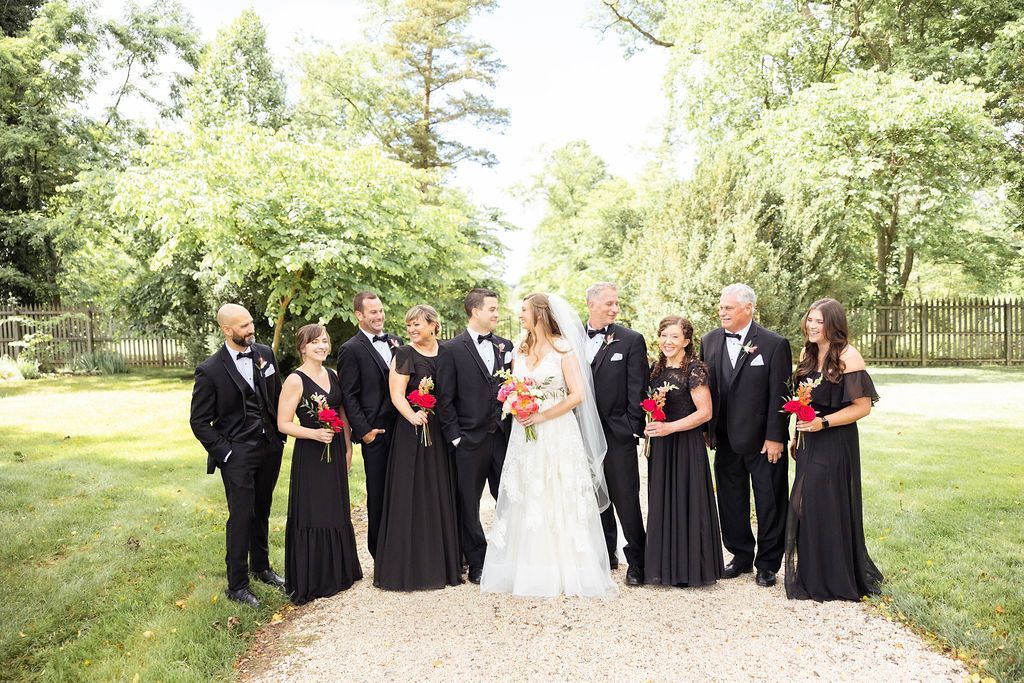 A wedding party stands in a grassy, tree-lined lane. A bride in a white gown is flanked by bridesmaids and groomsmen.