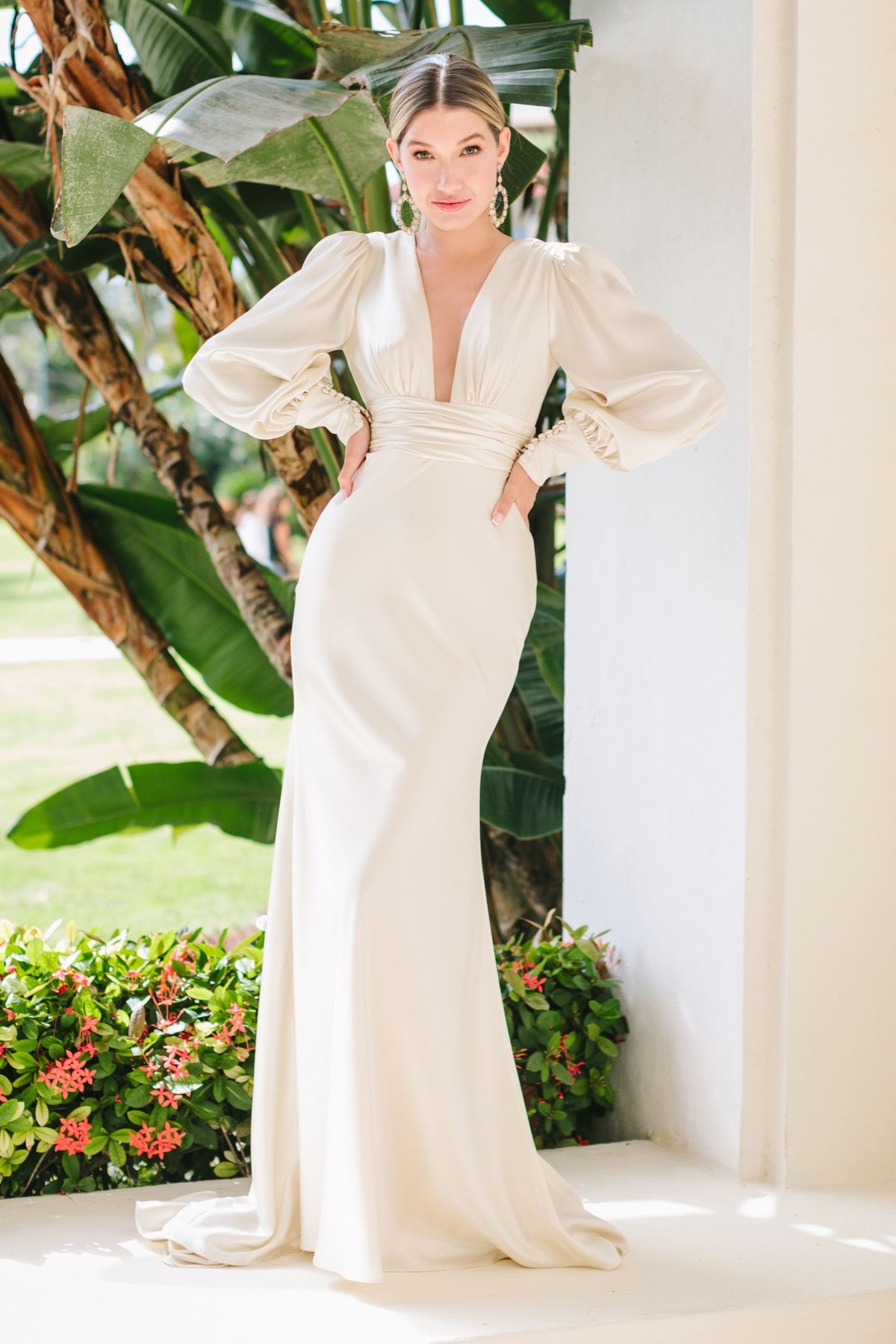 A person in a cream-colored, long-sleeved, floor-length gown with a V-neckline stands outdoors against lush greenery.