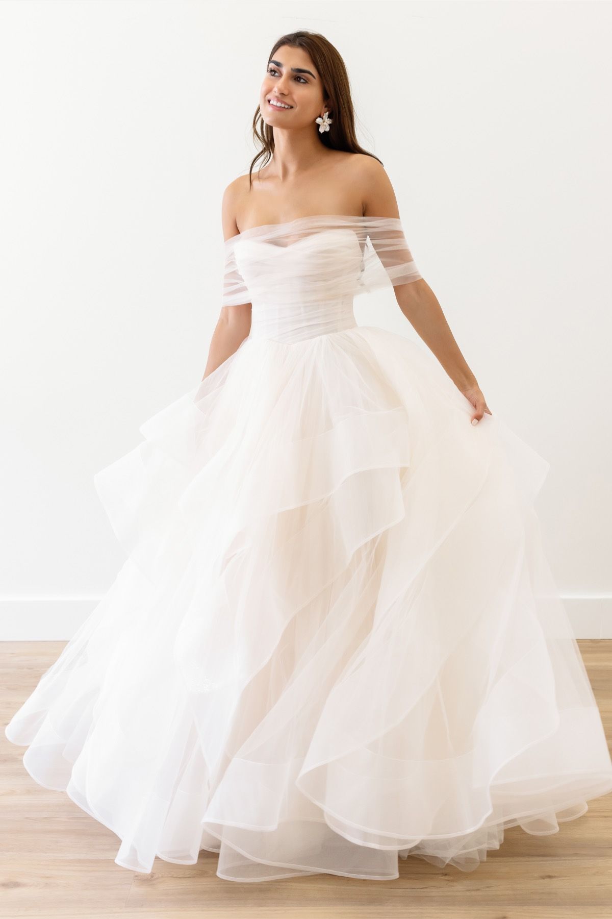 A person stands smiling in an off-the-shoulder, white layered ballgown wedding dress against a plain white background.