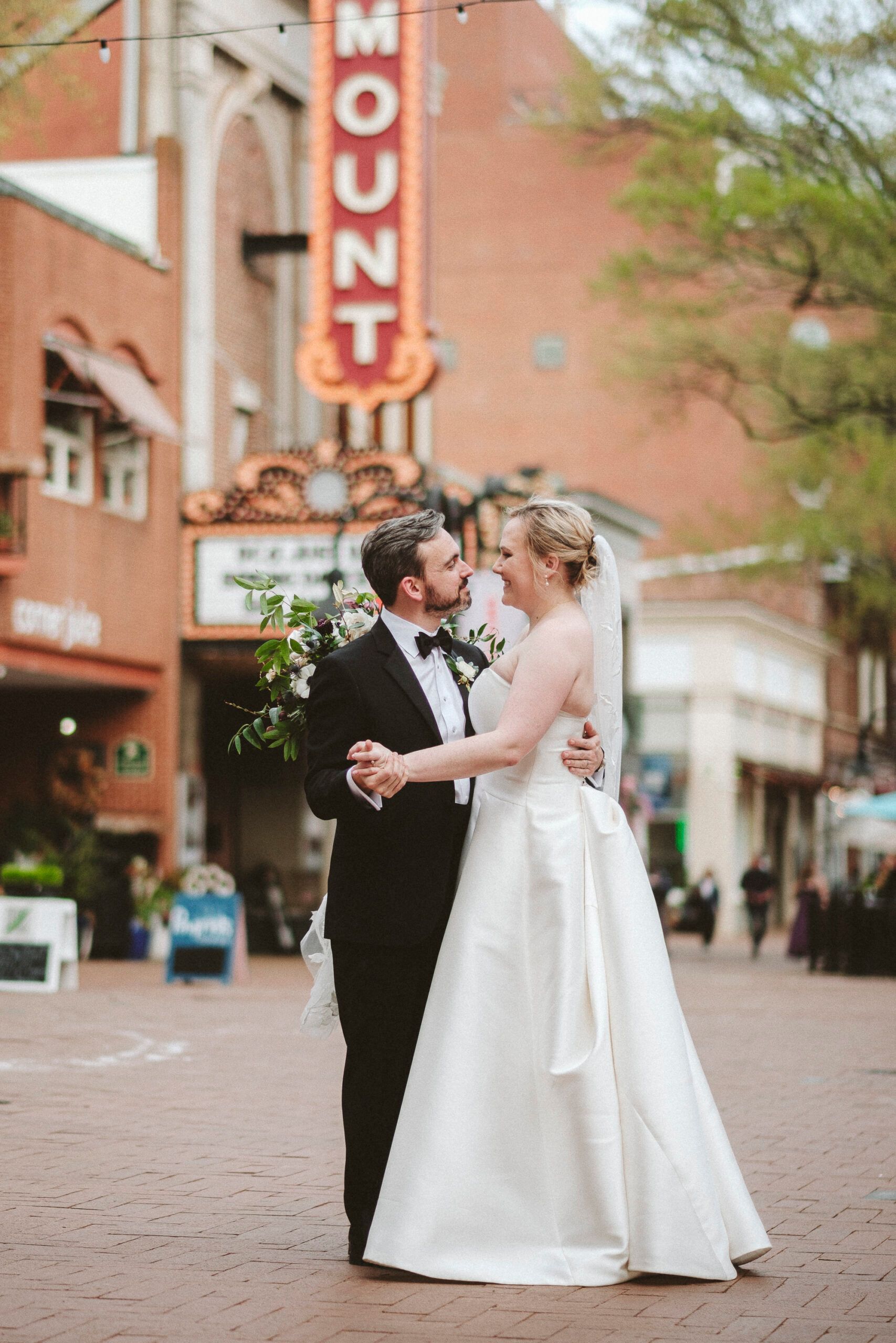 A couple in wedding attire dancing on a city street in front of a theater sign that says 