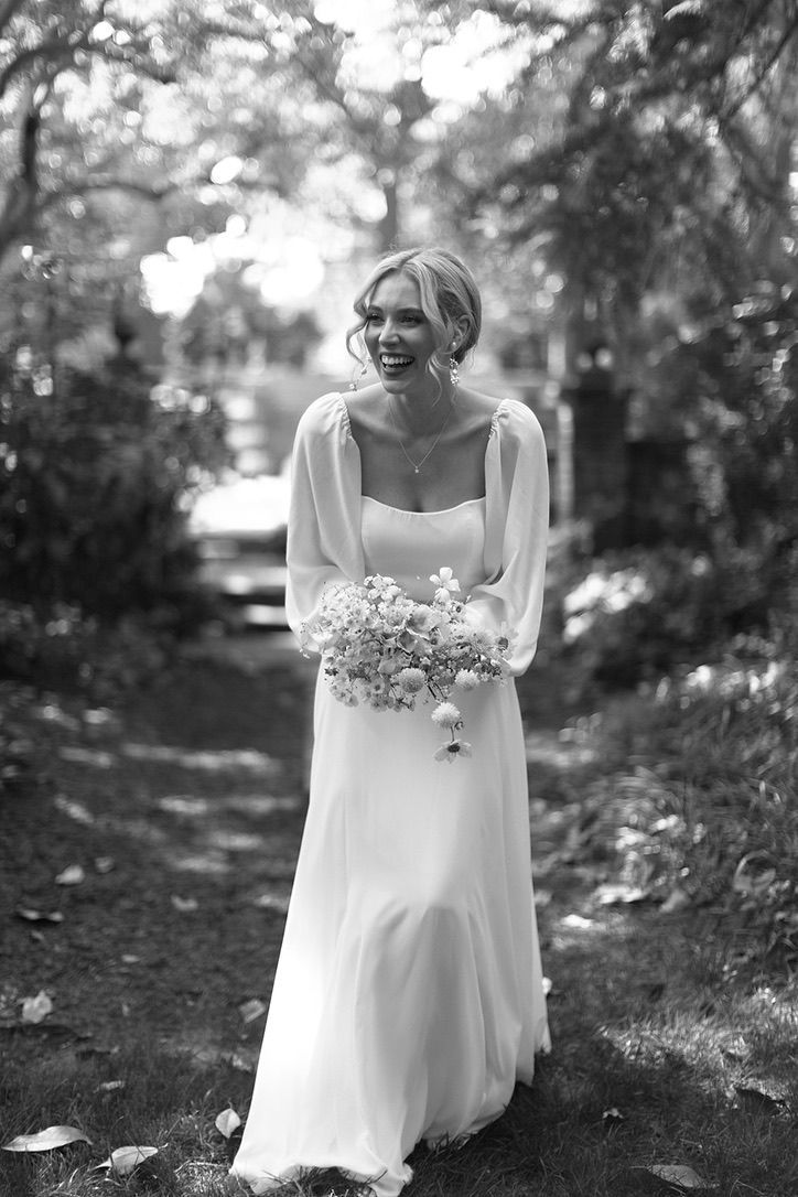 A person wearing a long-sleeved white wedding dress and holding a bouquet, laughing while walking through a garden.