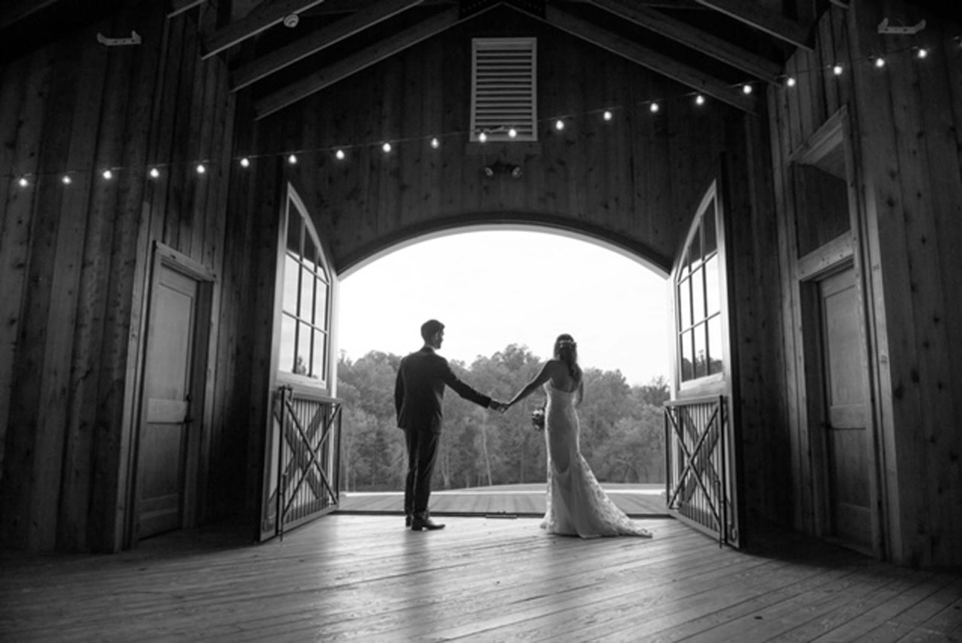 A couple stands hand-in-hand in the doorway of a rustic wooden barn, silhouetted against a bright, scenic outdoor view.