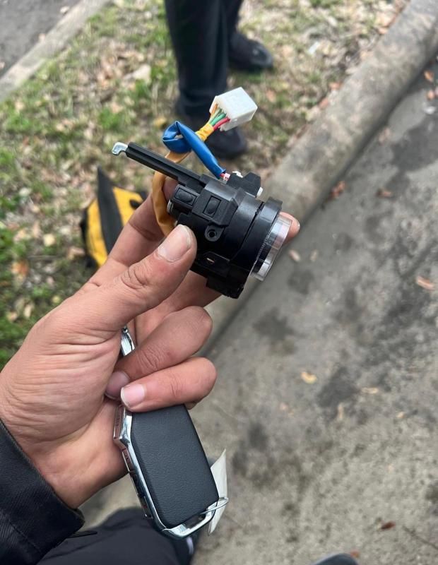Hand holding a small black camera or sensor module with wires and a connector outdoors near a curb