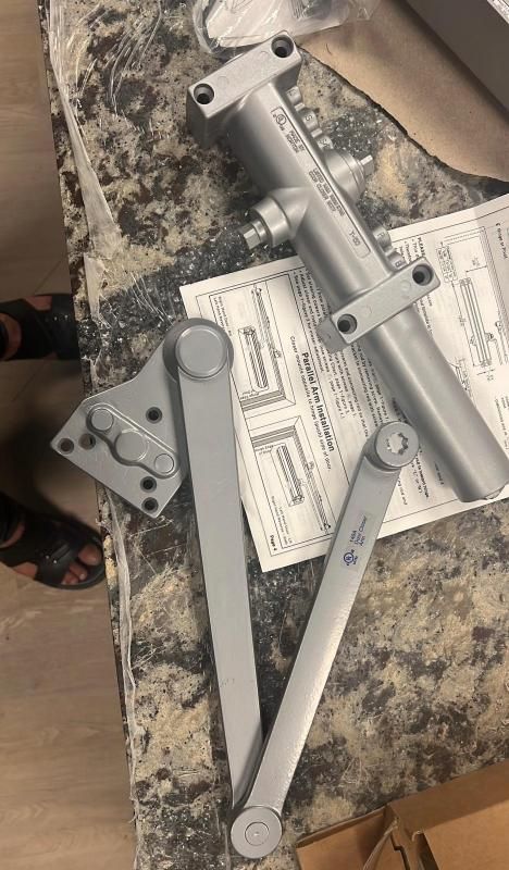 Disassembled silver metal door closer parts and brackets on a granite countertop with paperwork.