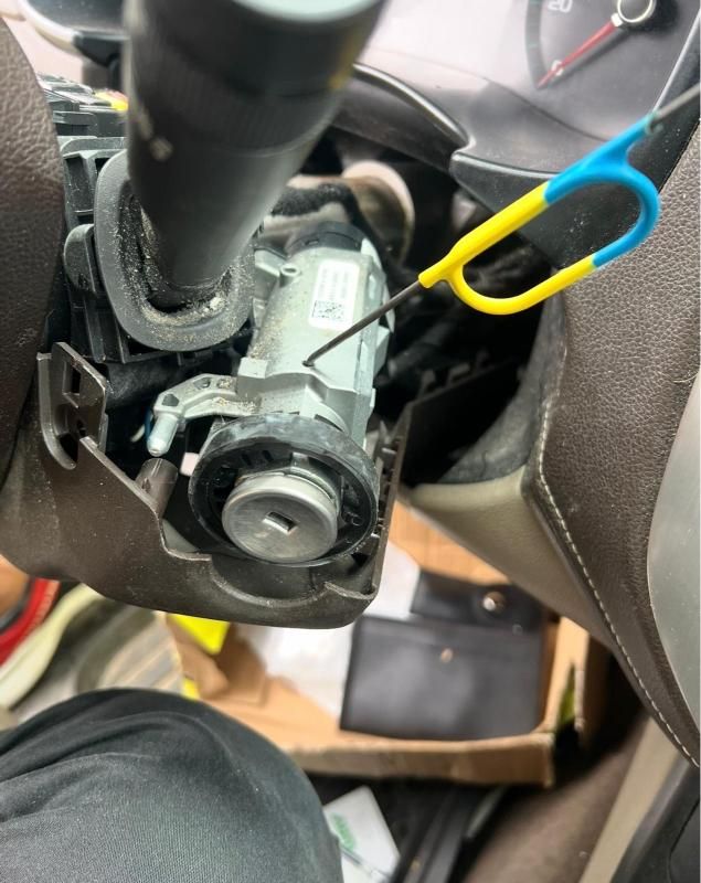 Close-up of a car steering column with exposed ignition parts and a blue-yellow tool inserted nearby