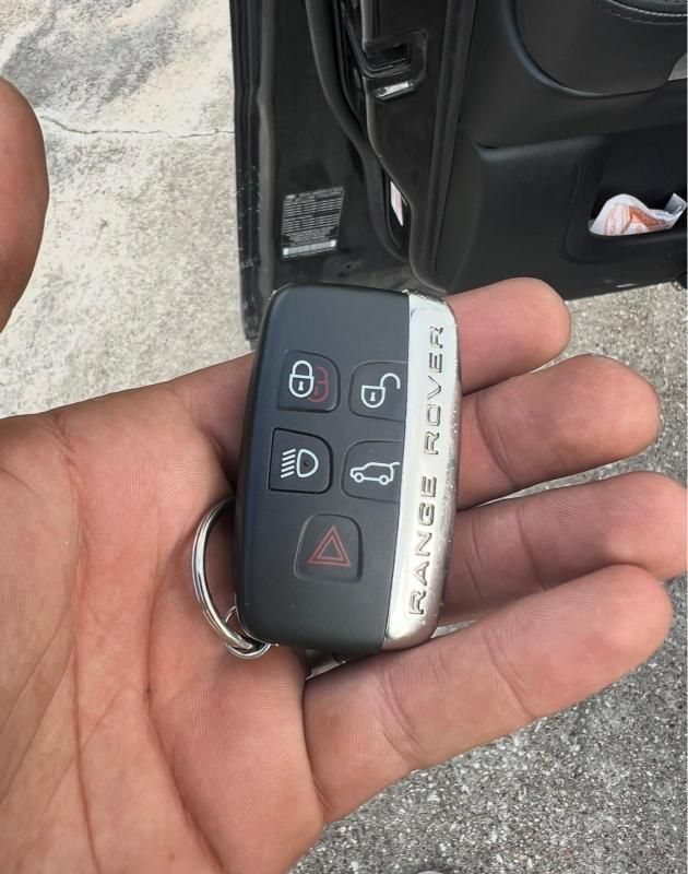 Hand holding a black car key fob with lock, unlock, trunk, and panic buttons beside an open car door.