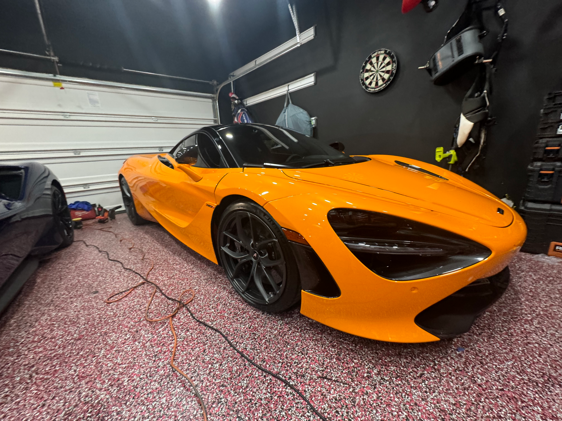 An orange sports car is parked in a garage next to a dart board.