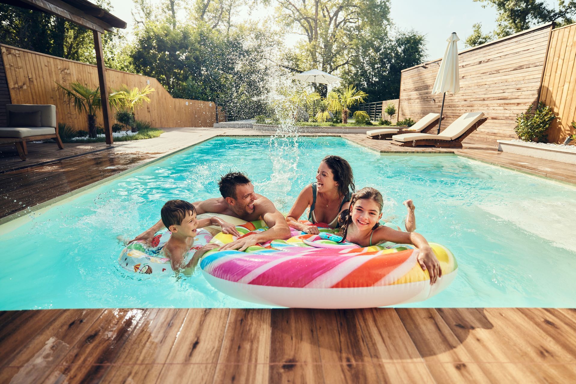 Family of four in a pool on an inflatable, laughing and splashing water. Sunny outdoor setting.