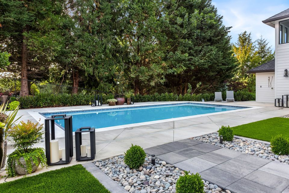 Rectangular pool in a backyard with greenery, gray stone patio, and a white house.