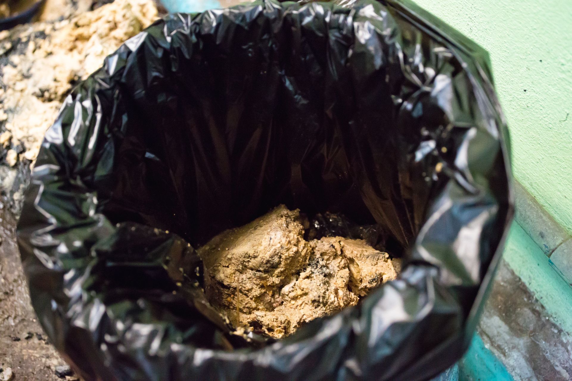 Black trash bin containing a pile of dirt or debris near a wall and floor.