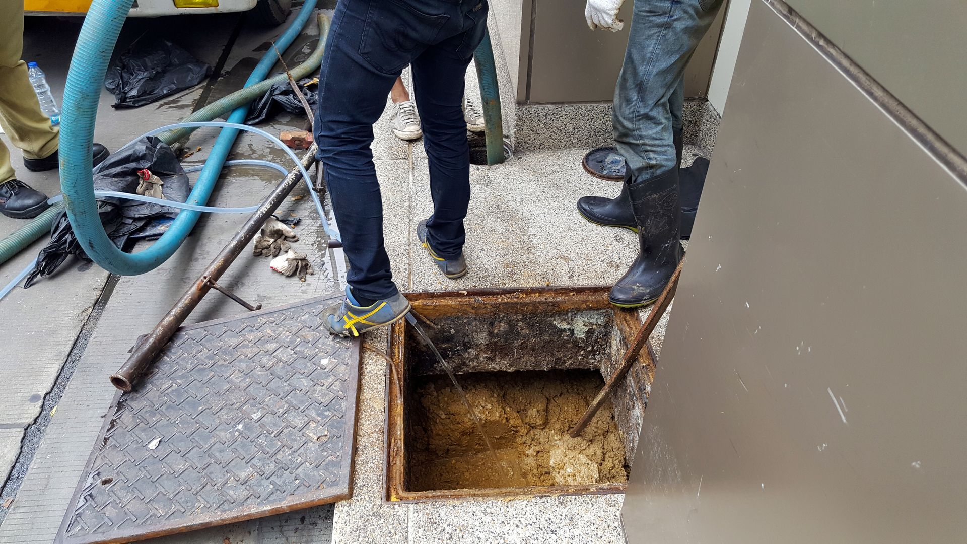 Workers standing beside an open ground hatch with hoses and tools on a wet construction site