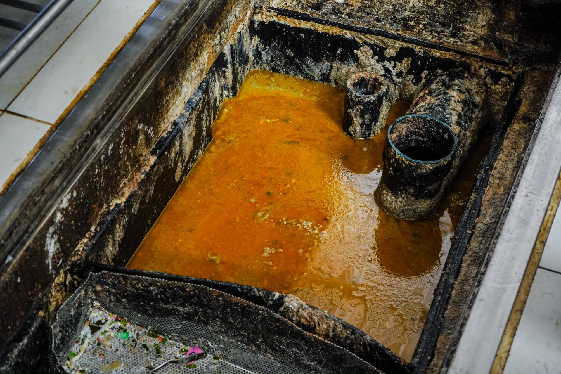 Orange liquid pooling in a grimy industrial tank or trough with black pipes and tiled edges