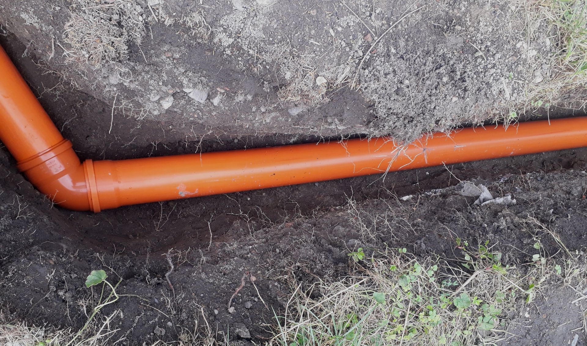 Orange drainage pipe running along a rocky soil embankment with sparse grass.