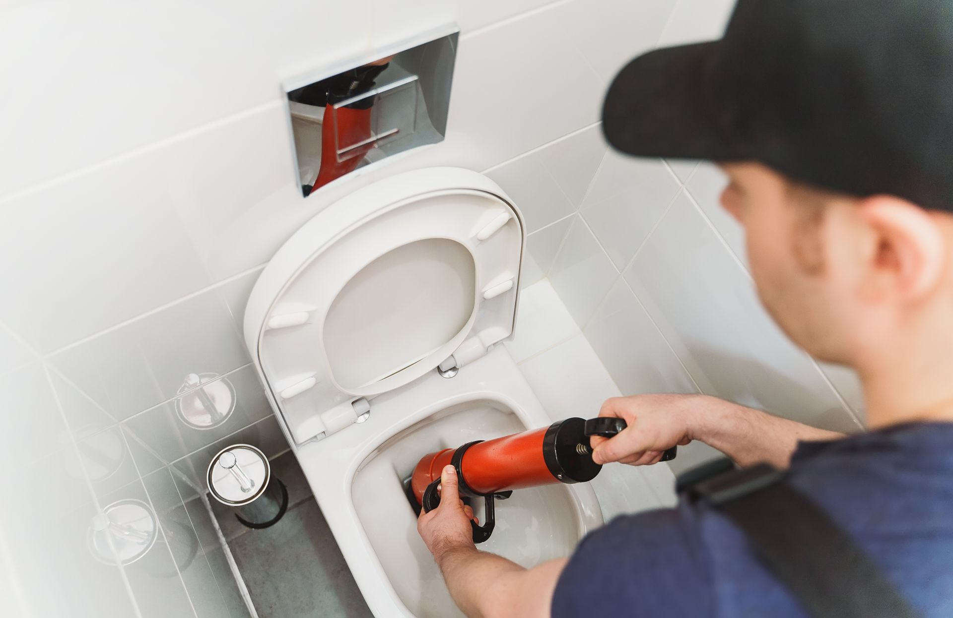 Plumber using a drill to work on a toilet in a white-tiled bathroom