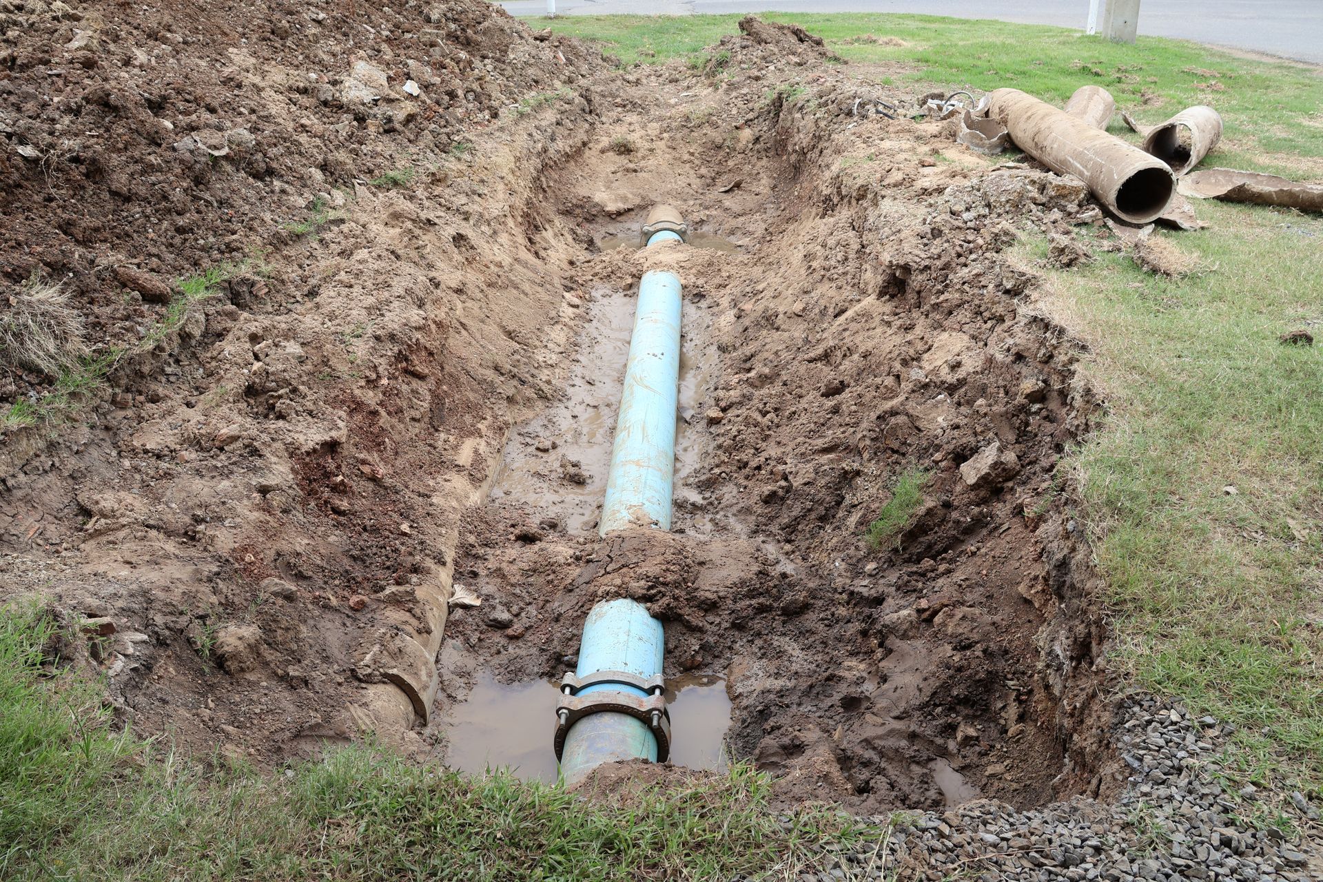 Exposed blue PVC pipe in a muddy trench, with a pipe section lying on grass nearby.