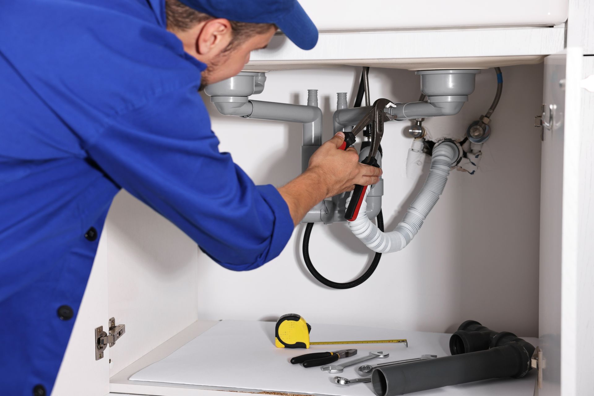 Plumber in blue uniform repairing under-sink pipes with a wrench in a kitchen cabinet