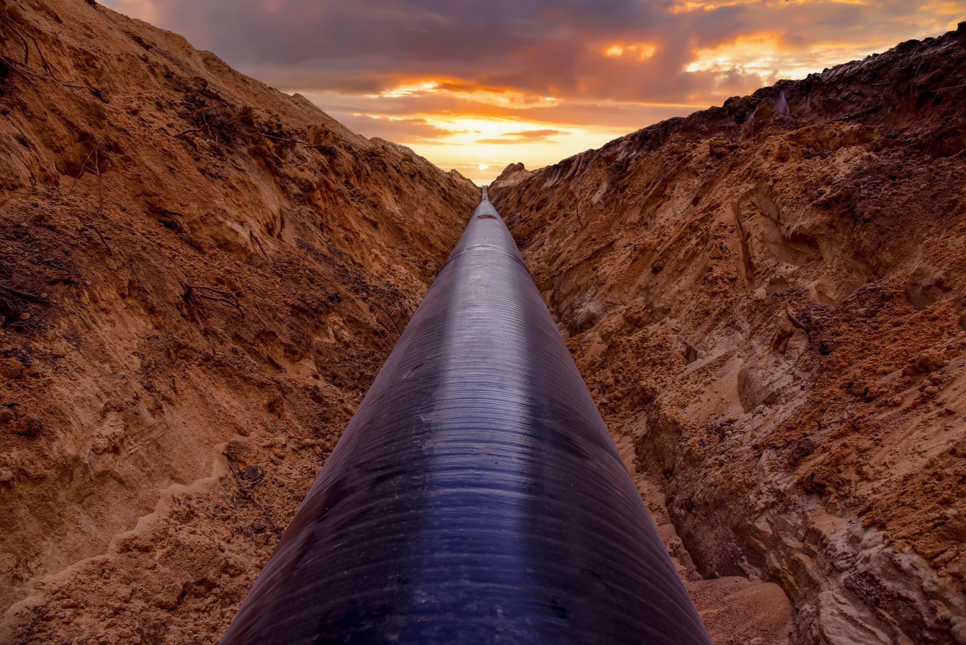 A black pipeline runs through a dry rocky trench at sunset under an orange sky.