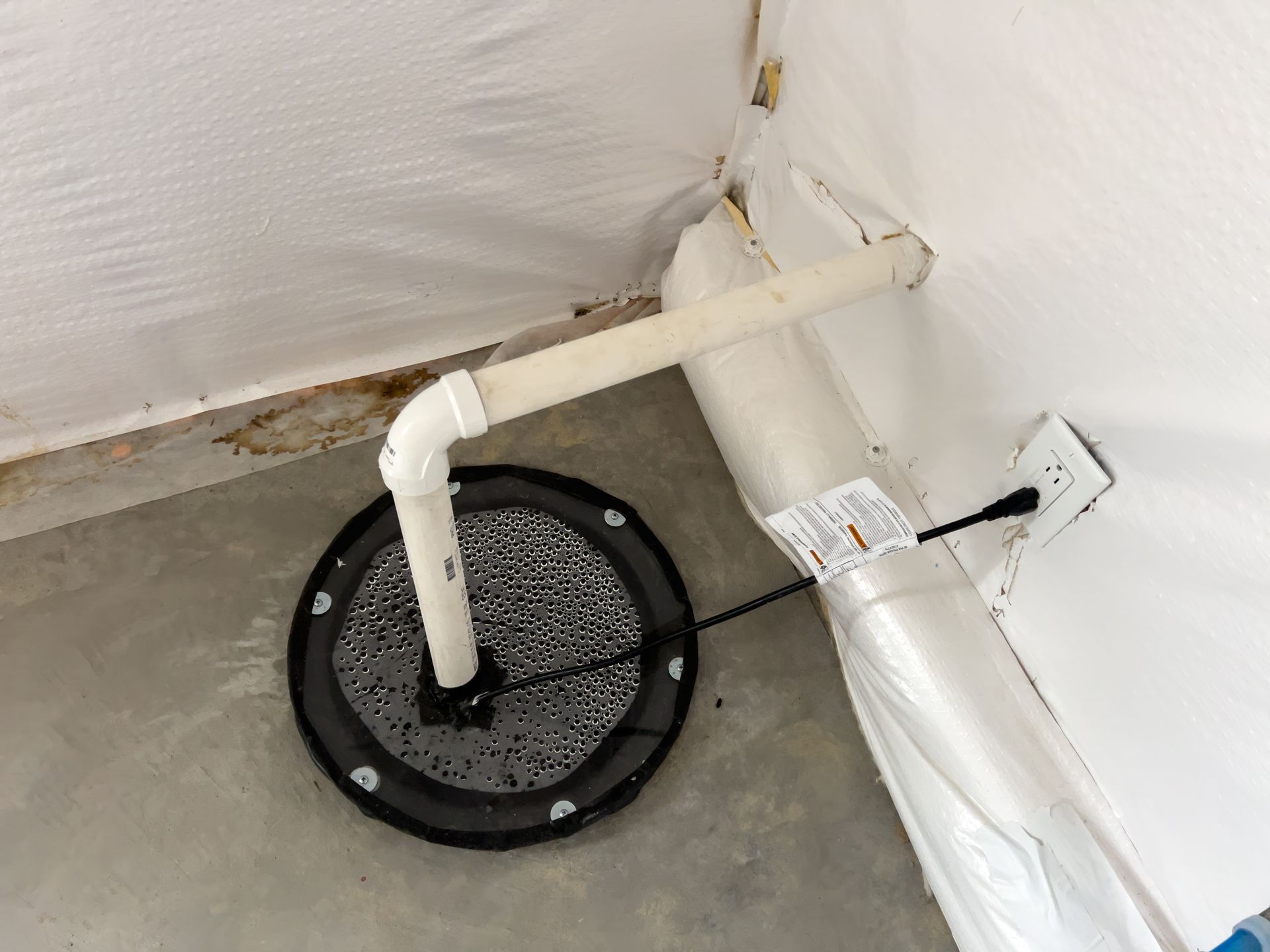 Basement corner with white PVC pipe connected to a black floor drain on a concrete floor.