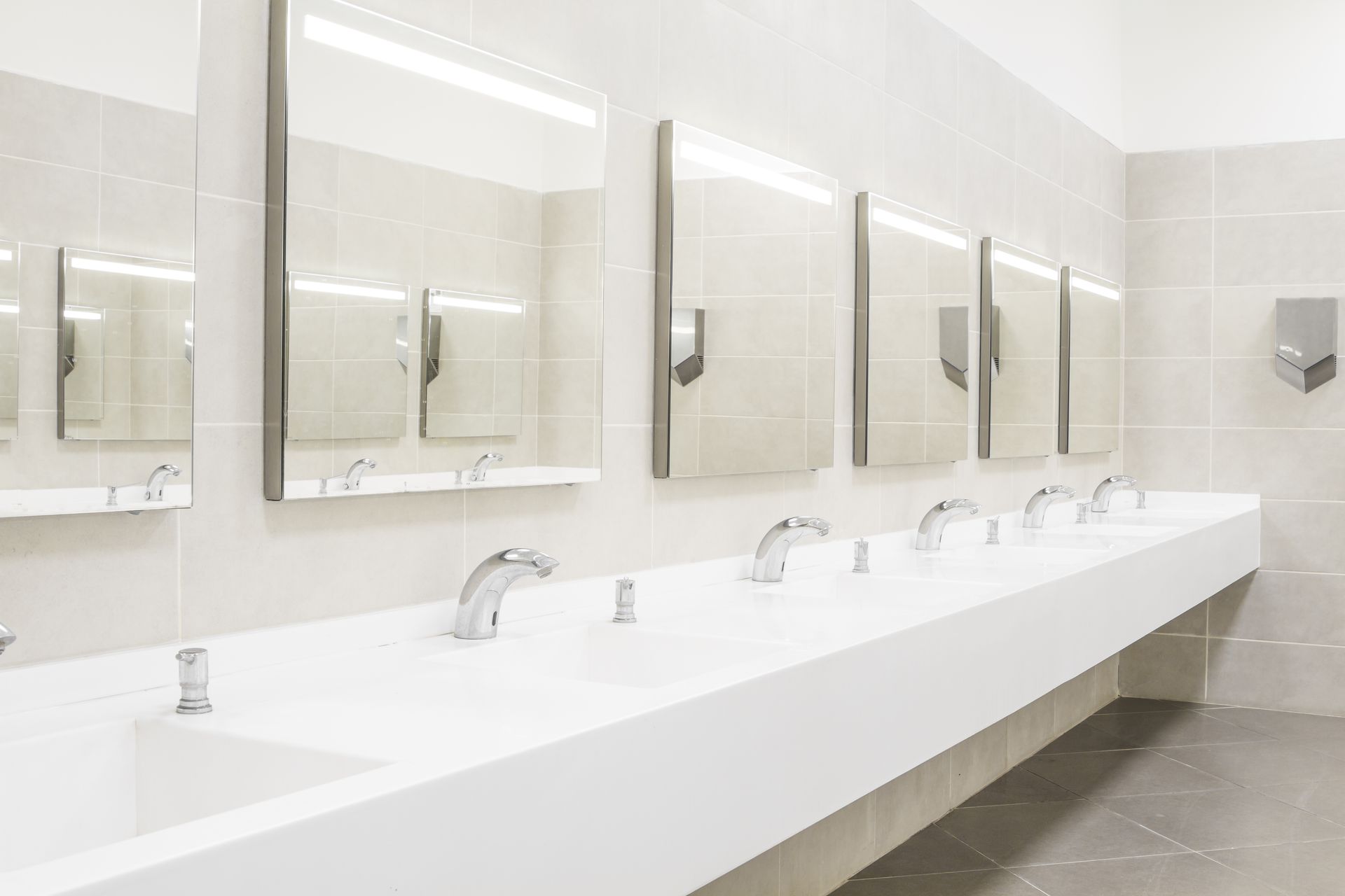 Bright modern public restroom with a long white sink counter and mirrors on tiled walls