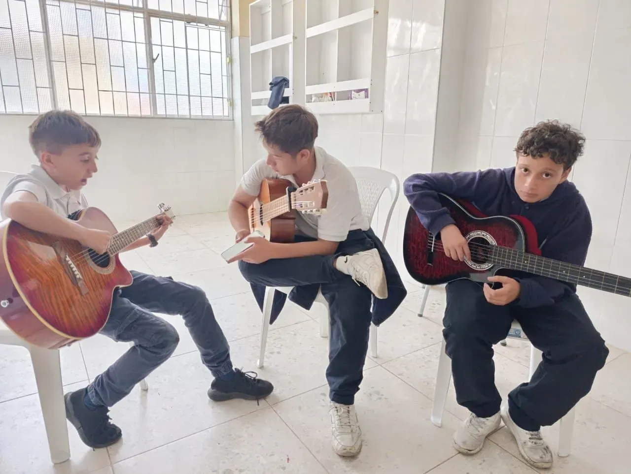 Tres niños sentados en el interior, cada uno tocando una guitarra acústica en una habitación luminosa.