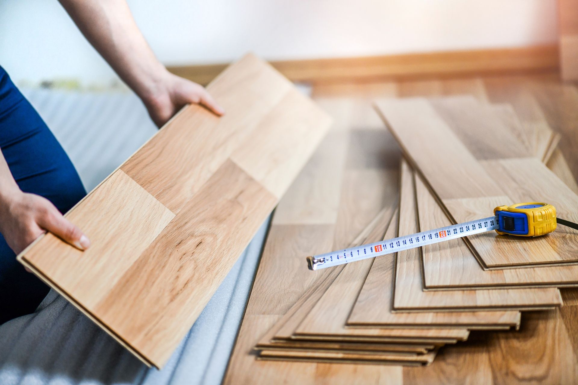 Hands holding wooden flooring planks beside a tape measure and stacked boards on a floor