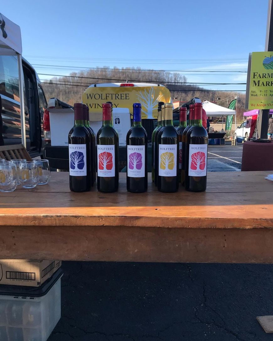 Six bottles of wine on a wooden market table outdoors, with a sunny street and vendor tents in the background