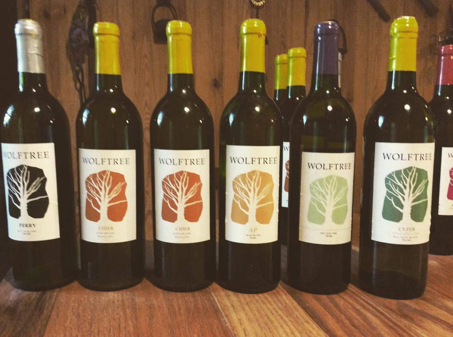 Six wine bottles lined up on a wooden table, with white, orange, yellow, and green labels.