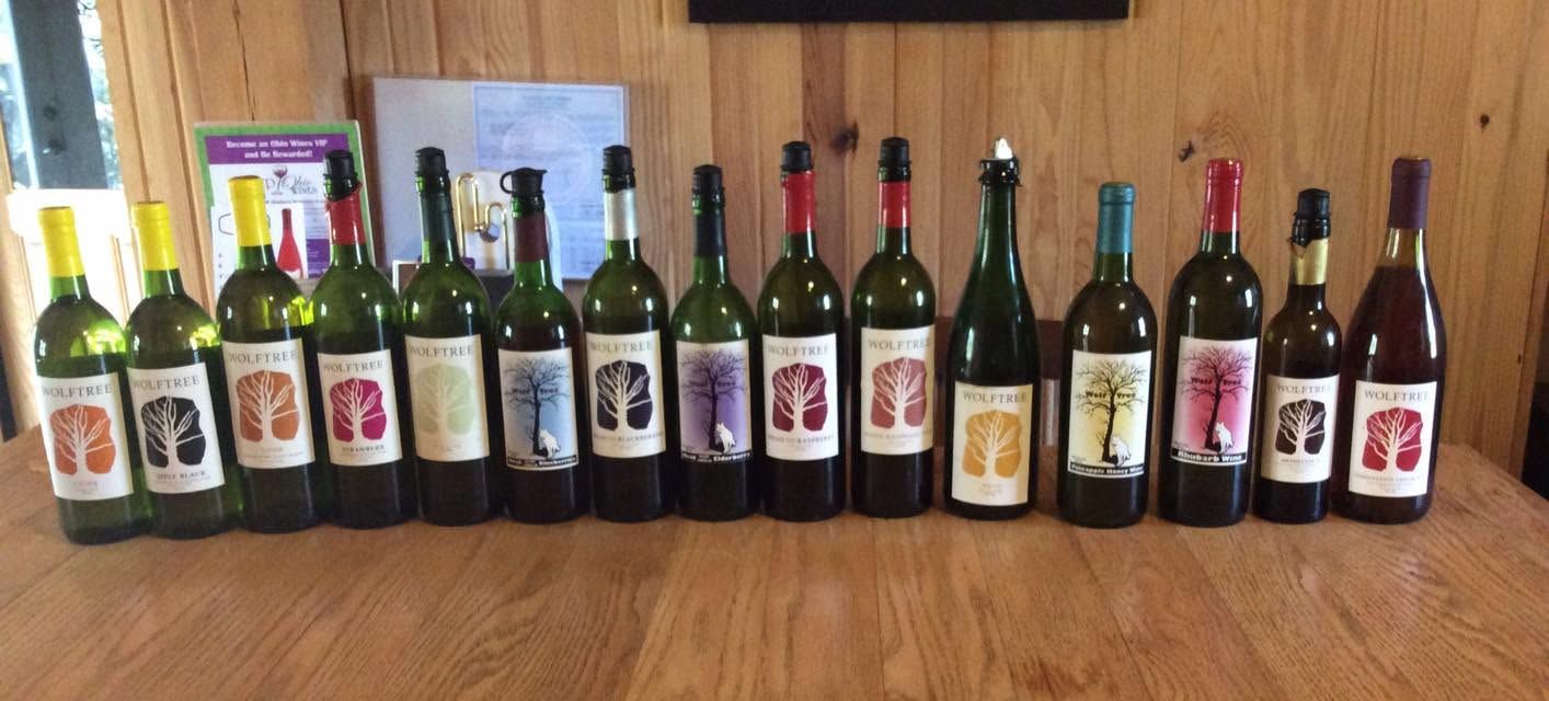 Assorted wine bottles lined up on a wooden table in a room with white cabinets.