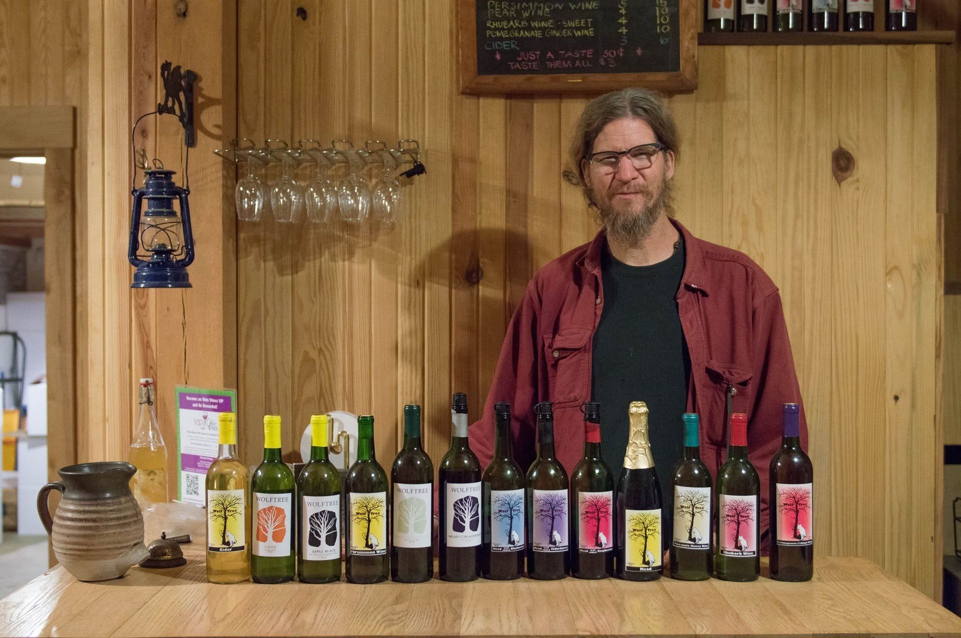 Man standing behind a counter lined with assorted beer bottles in a wood-paneled bar.