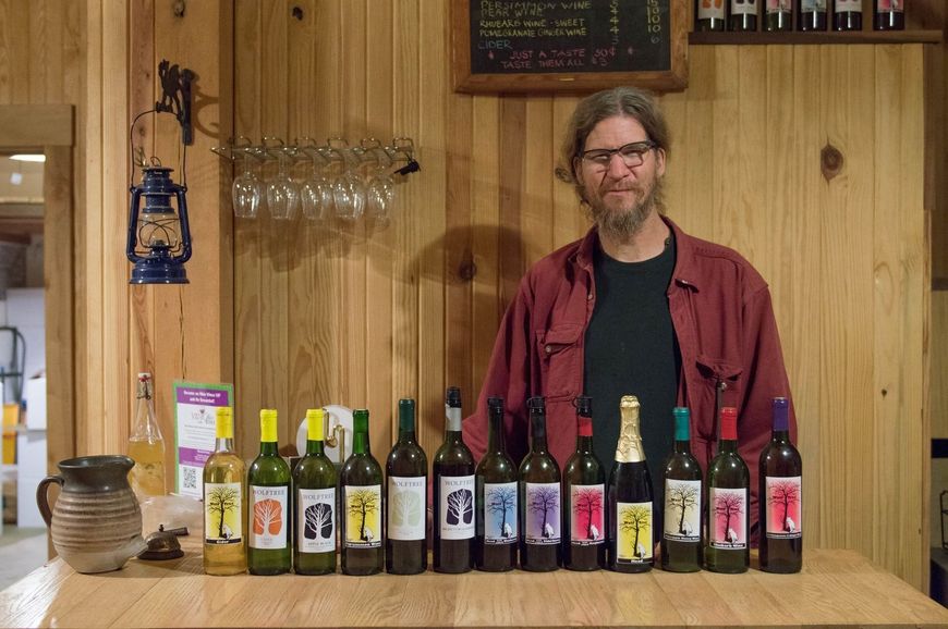Man standing behind a counter lined with assorted beer bottles in a wood-paneled bar.
