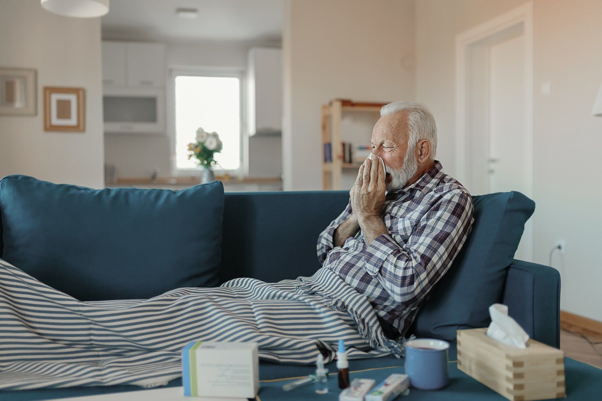 An elderly person sits on a couch, sneezing into their hand. They are covered by a striped blanket in a living room.