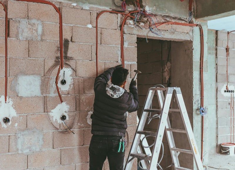 Une personne effectue des travaux de câblage sur un mur intérieur inachevé, à côté d'une échelle et d'une porte.