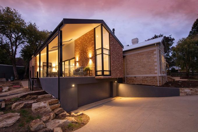 Maison moderne avec de grandes fenêtres éclairées au crépuscule, à côté d'un mur en pierre et d'une allée.